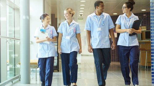 a group of four young trainee nurses including male and female nurses , walk towards camera down a hospital corridor . They are wearing uk nurse uniforms of trousers and tunics.